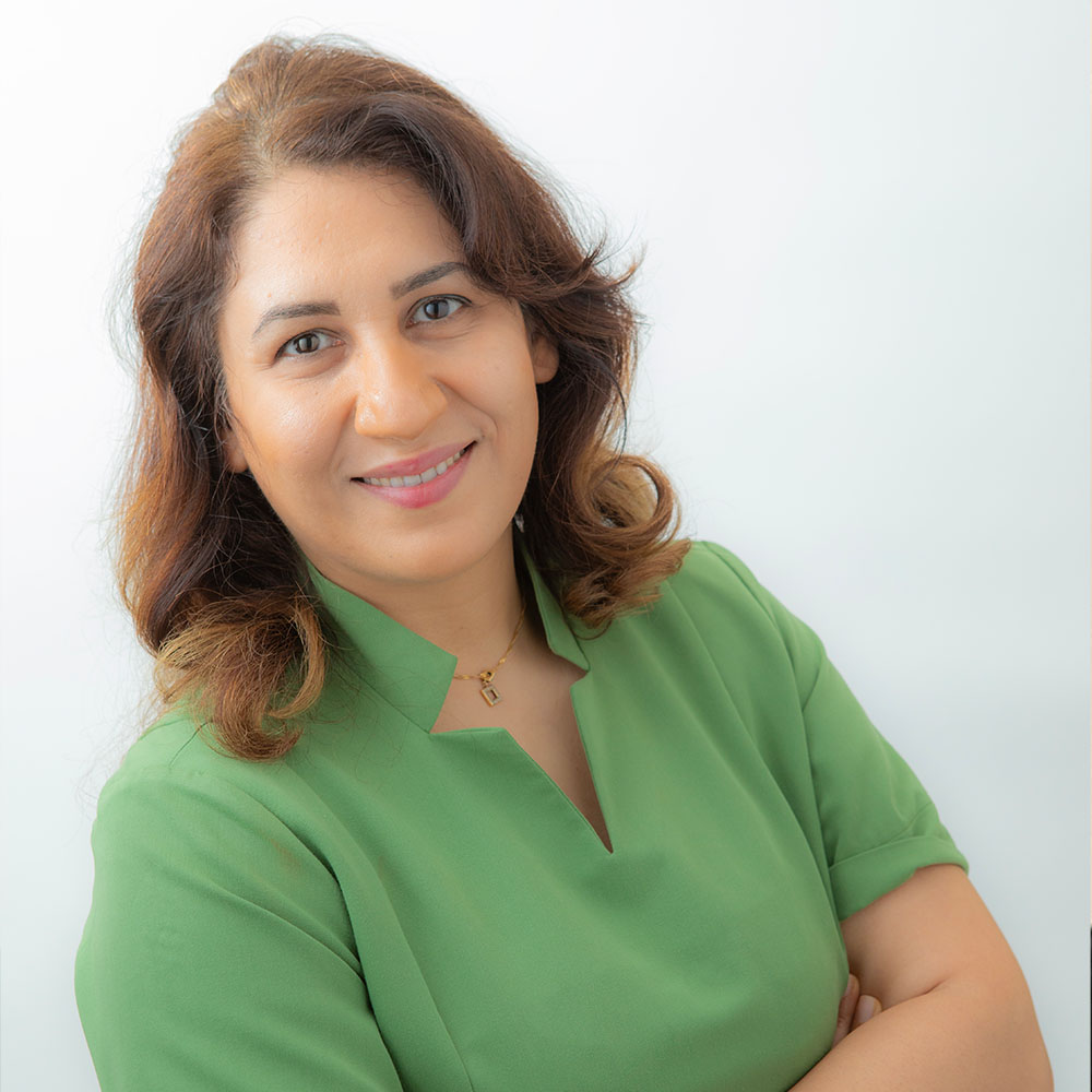 The image shows a woman wearing a green shirt, standing with her arms crossed, posing for a portrait-style photograph against a light background.