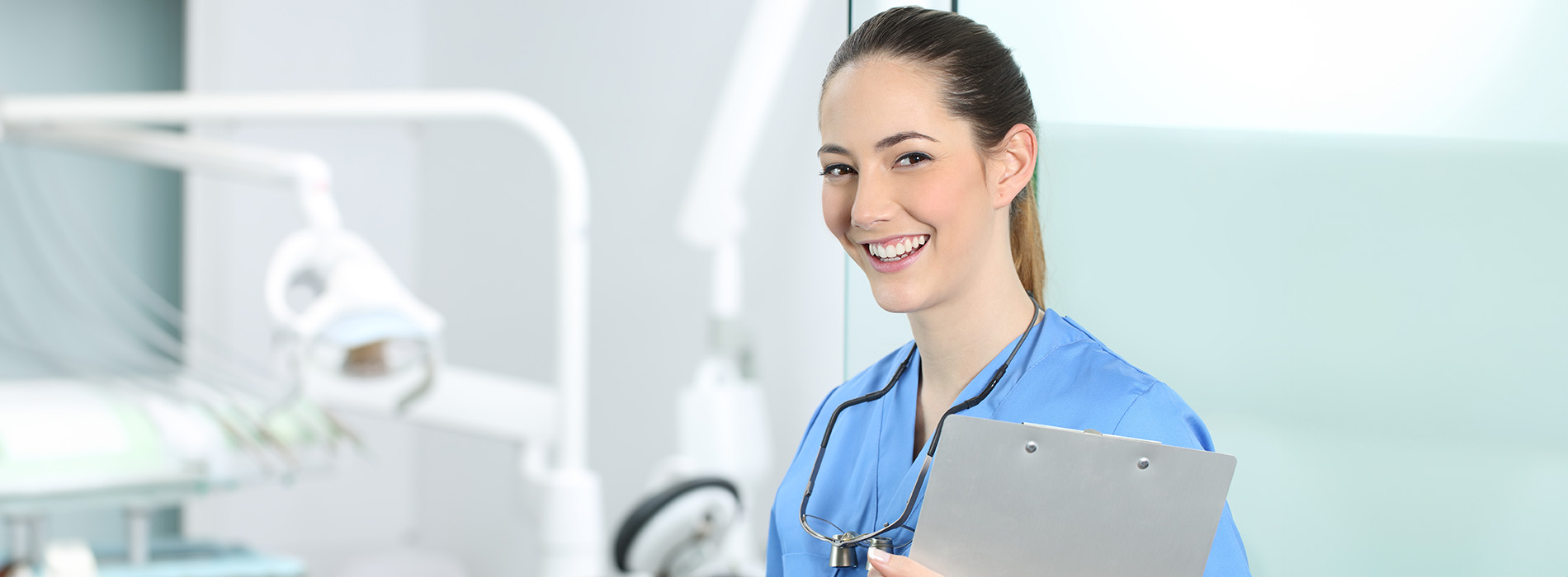 The image shows a woman in a blue scrub uniform standing in front of a dental chair, smiling at the camera with a tablet in her hand.