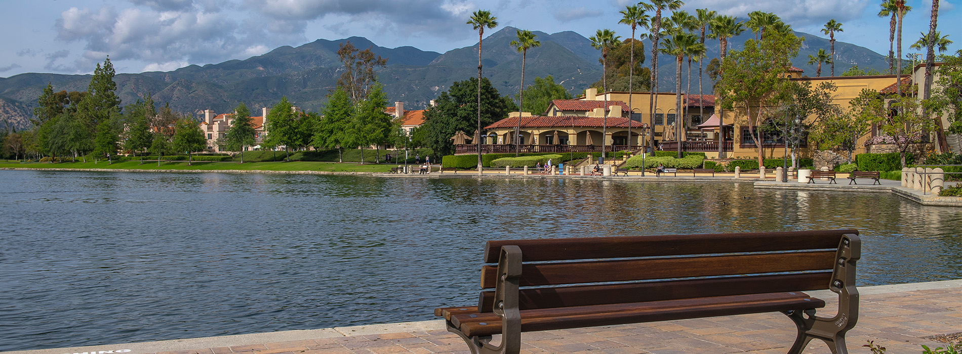 The image shows a serene riverside scene with a bench overlooking a calm body of water, surrounded by lush greenery and mountains in the distance.