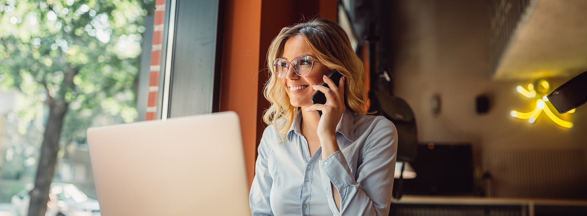 A woman wearing glasses is seated at a laptop, engaged in a phone call, with an indoor setting featuring a restaurant ambiance.