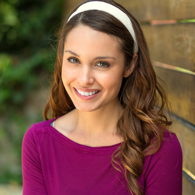 The image features a woman with long brown hair smiling at the camera, wearing a purple top, a white headband, and posing against a wooden fence.