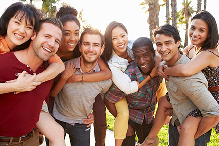 The image shows a group of people posing together outdoors during the daytime, with clear skies and sunlight illuminating them. They appear to be friends or family members embracing each other.