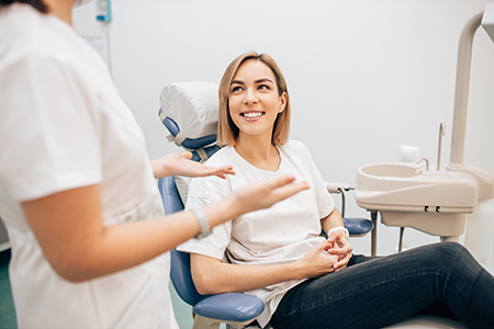 The image shows a woman sitting in a dental chair, smiling at the camera, with a dental professional standing nearby who appears to be engaged in conversation with her.